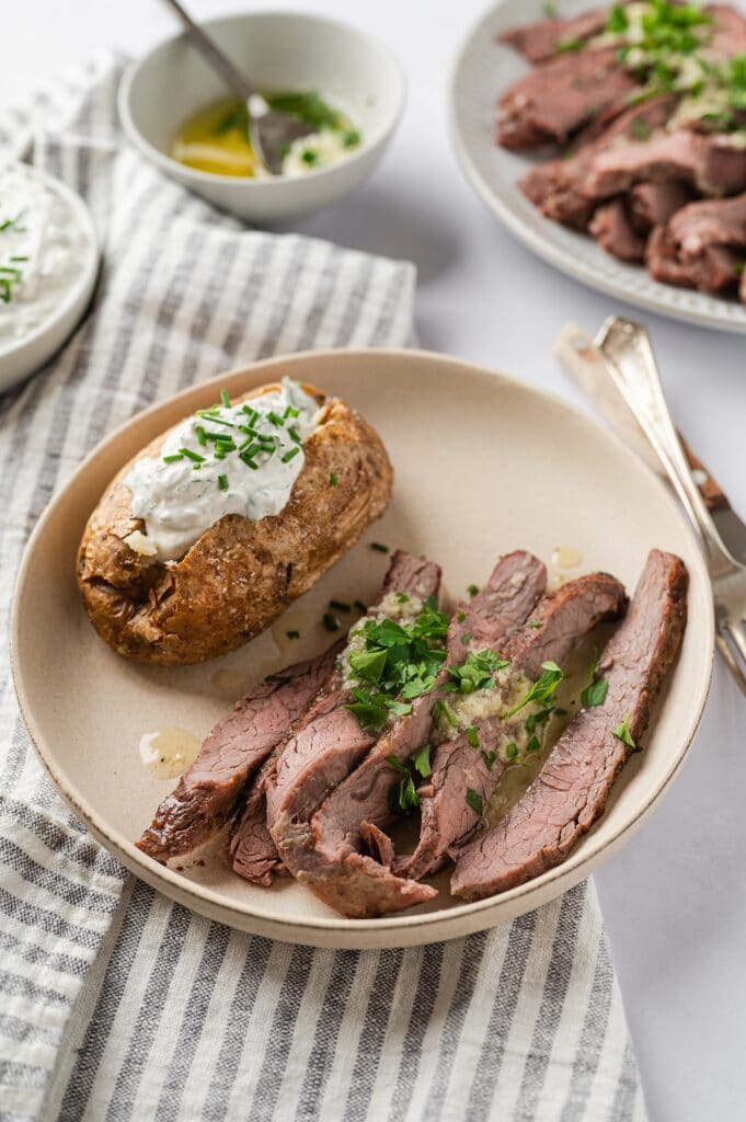 flank steak with garlic butter sauce and a baked potato with sour cream and chives on a plate