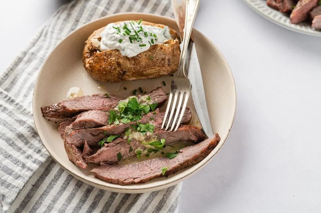 flank steak with garlic butter sauce on a plate with a baked potato with sour cream, chives and a fork