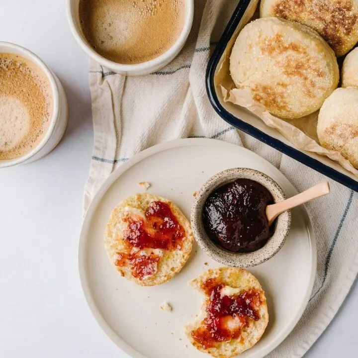 huckleberry english muffin with jam on a plate with jam and coffee and a tray of muffins in the background