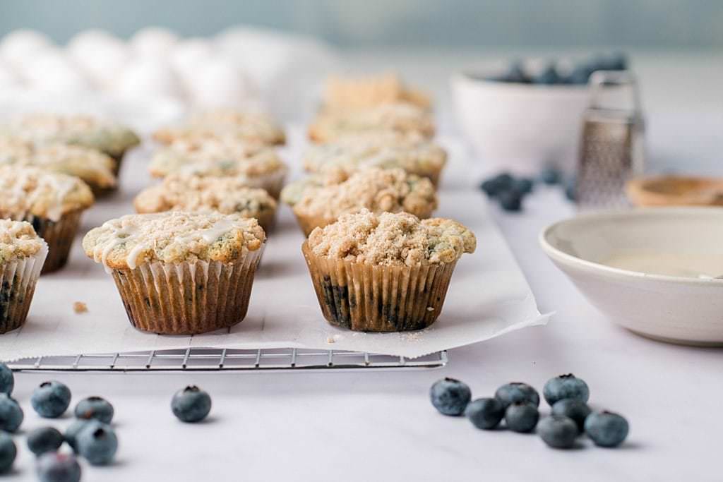 Blueberry muffins with crumb top on a wire rack with a bowl of glaze and blueberries