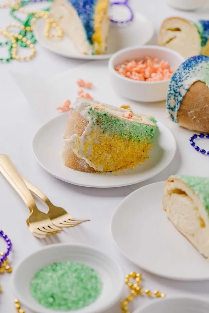 slice of homemade Mardi Gras cake on white plate with plastic babies in white bowl and two gold forks
