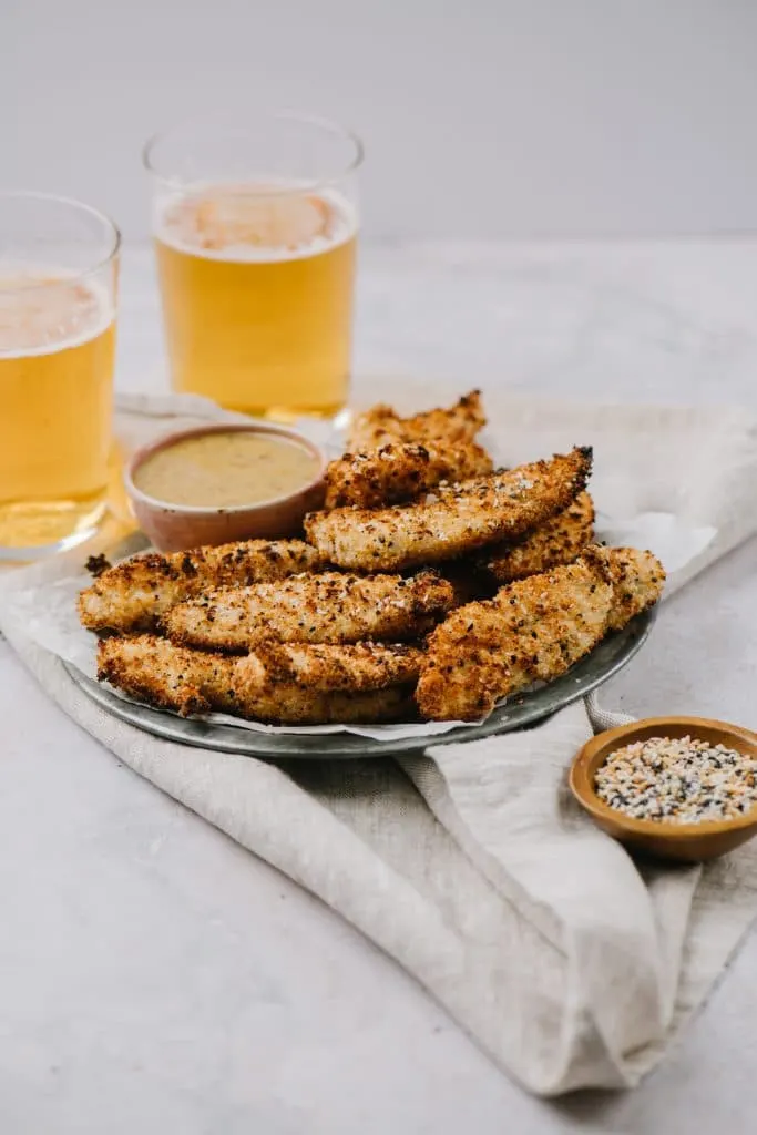 a bowl of everything seasoning next to a plate of air fryer chicken fingers with two beers next to it