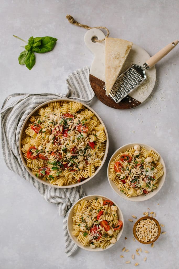 fresh tomato and basil pasta in large white bowl with serving spoon and two small bowls with fresh basil leaves and block of parmesan cheese