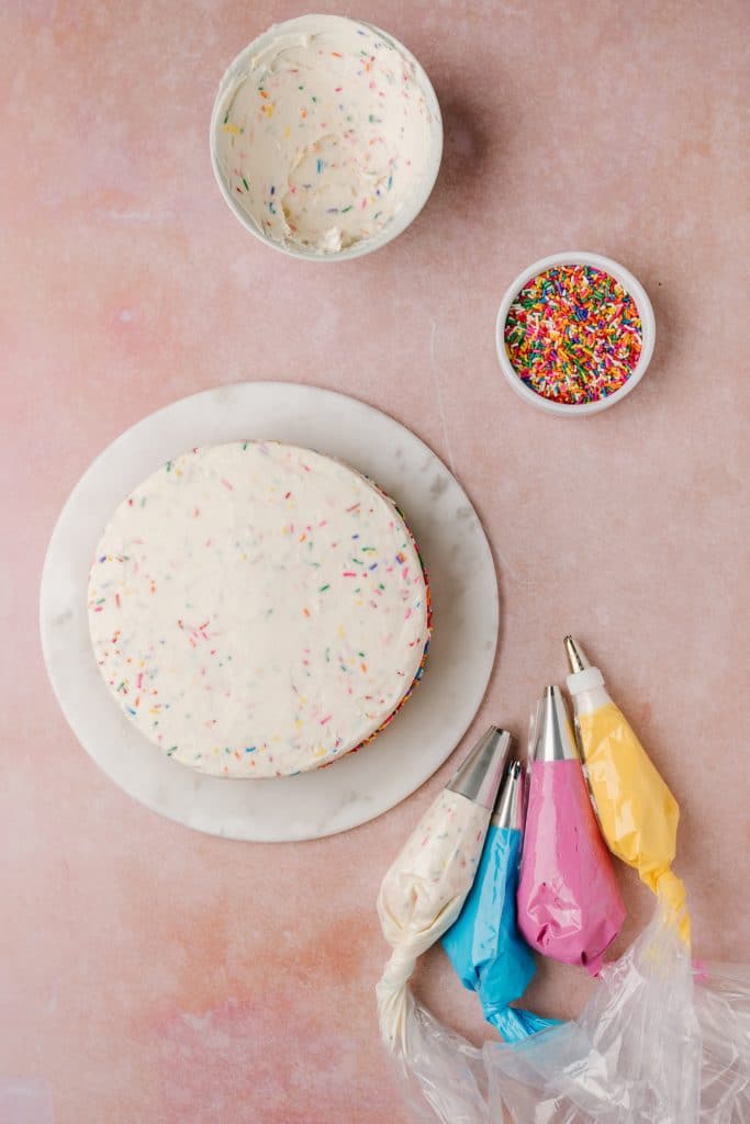 iced vanilla cake with white, blue, pink, yellow buttercream in pastry bags and rainbow sprinkles in bowl
