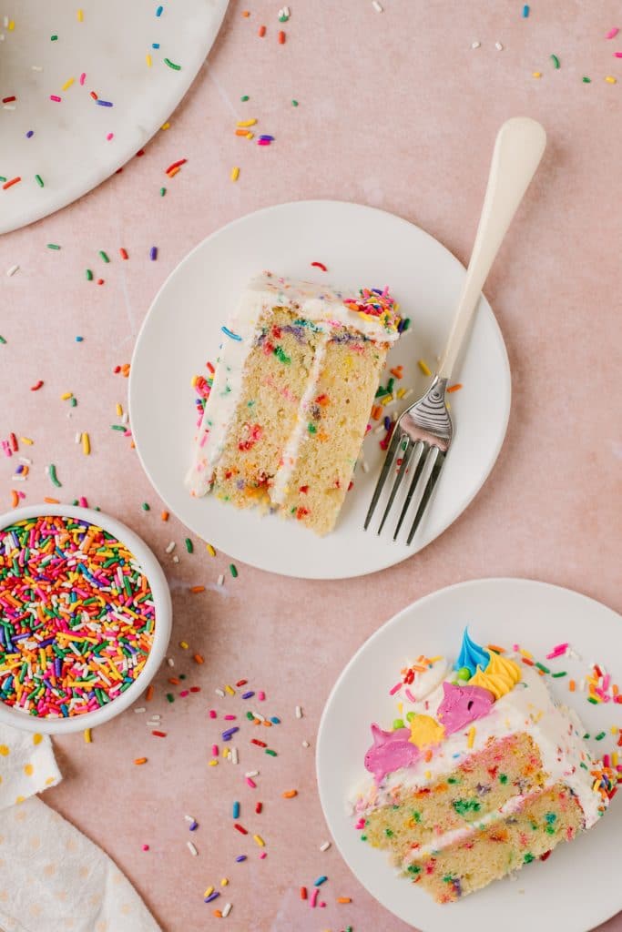 slices of vanilla cake with rainbow sprinkles on white plates with fork and sprinkles on side