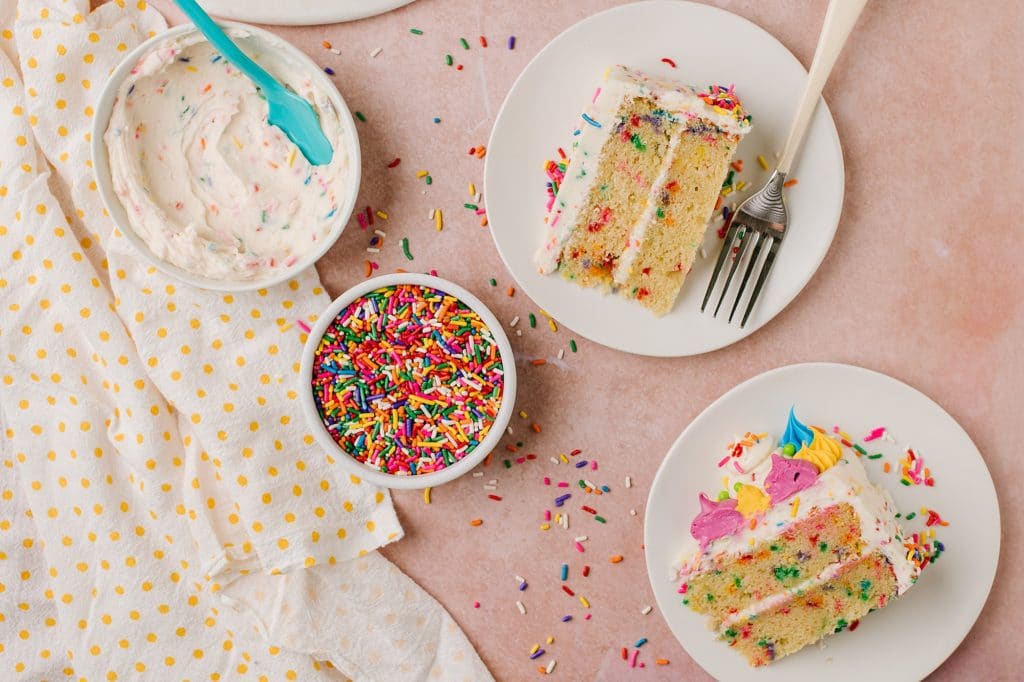 two homemade confetti cake slices with a bowl of funfetti buttercream and a bowl of sprinkles