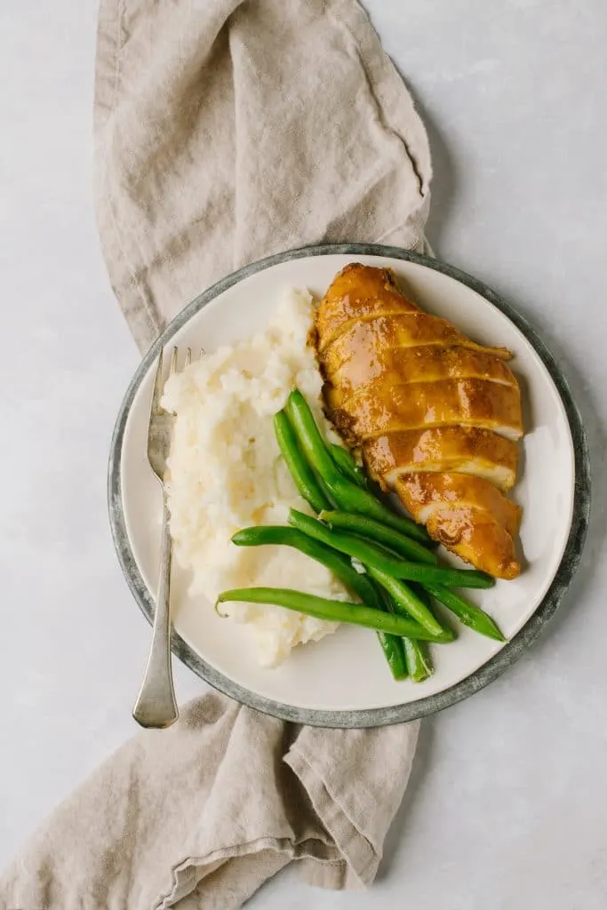sliced chicken with sauce with mashed potatoes and green beans on white plate with gray napkin and fork