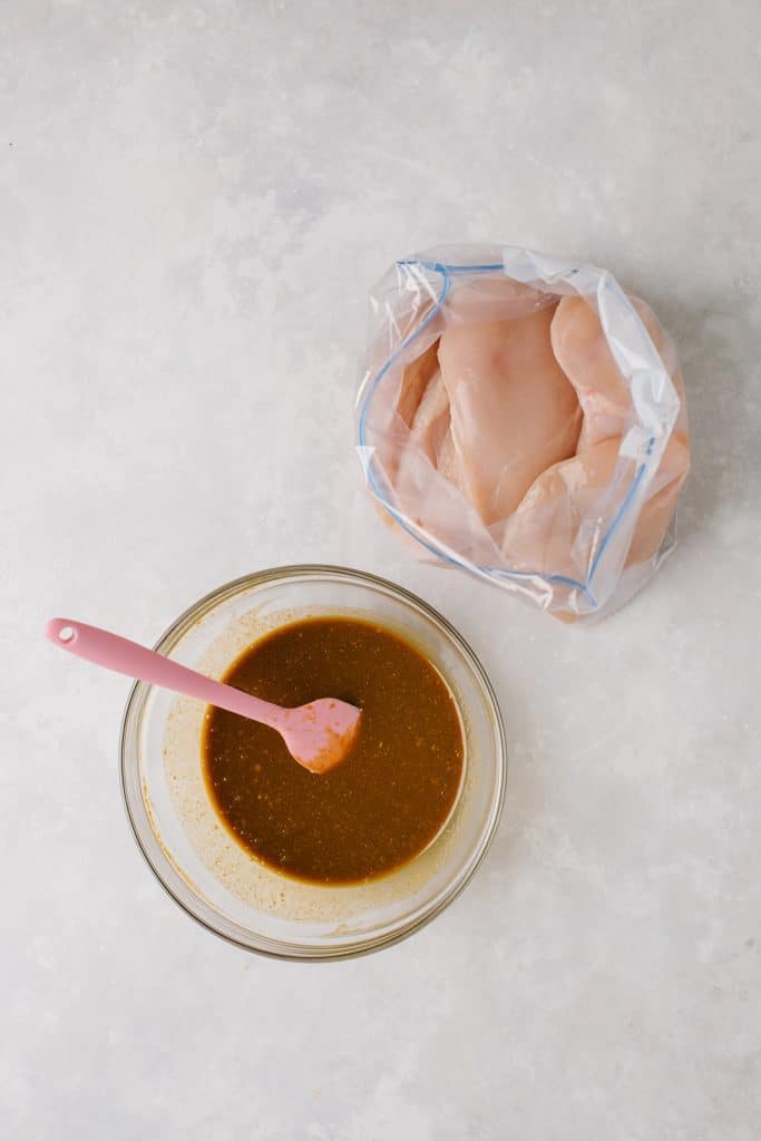 marinade in glass bowl with pink spatula, chicken breasts in plastic bag