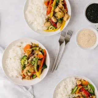 bowls of healthier orange chicken and vegetables with two forks and two bowls of sesame seeds