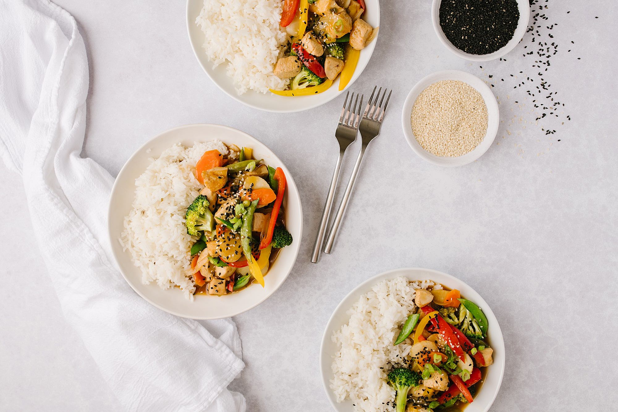 three bowls of healthier chicken and vegetables and rice with two forks and two side bowls of black and white sesame seeds