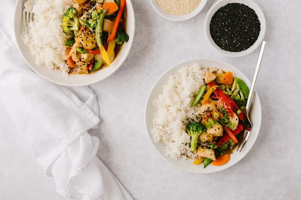 two bowls of healthier chicken and vegetables and rice with two forks and two side bowls of black and white sesame seeds