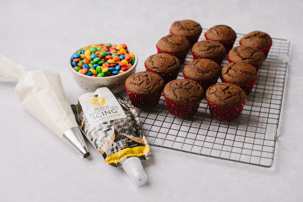 chocolate cupcakes on wire rack with pastry bag filled with frosting and bowl of m&ms
