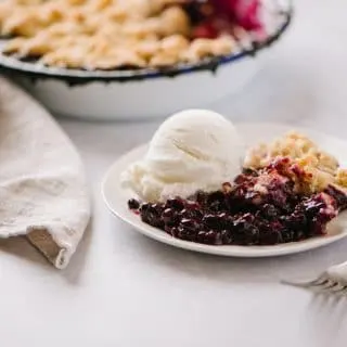 plate of the best blueberry crisp with a scoop of vanilla ice cream on a plate with a fork and linen napkin with a pan of crisp in the background