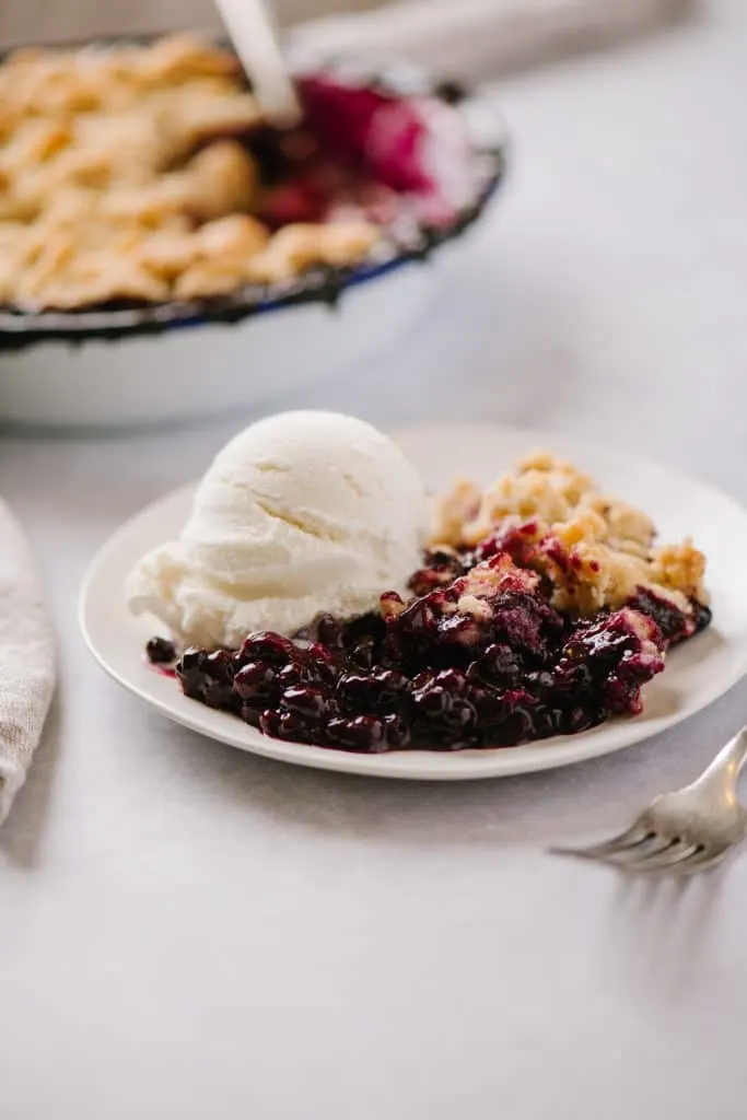 side angle shot of blueberry crisp and vanilla ice cream on a plate with a pan of crisp in the background
