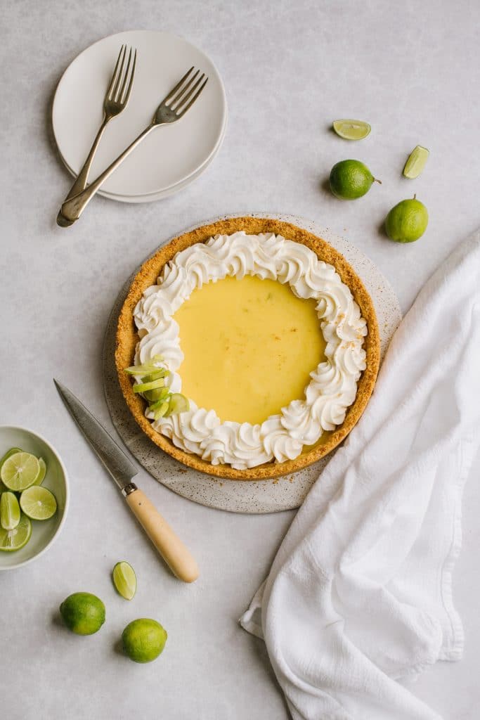 lime curd pie topped with whipped cream with fresh limes, plates and forks on the side