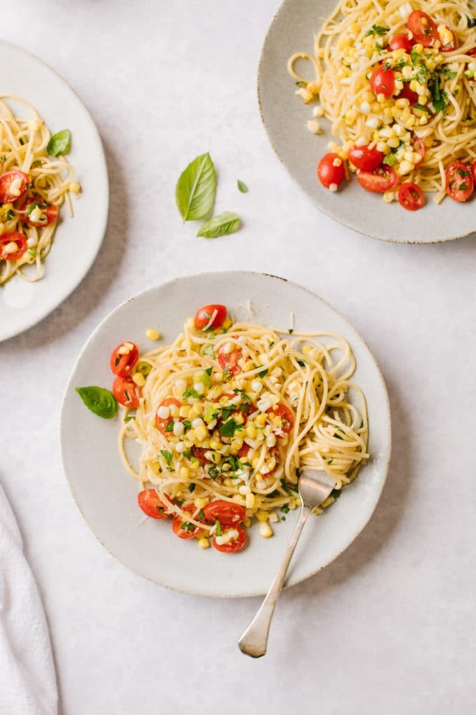 a plate of pasta with corn gremolata and a fork with pasta twisted around it and two plates out of the shot with basil leaves