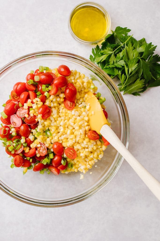 a bowl of corn, tomatoes, and green onions with a small bowl of olive oil and parsley next to it