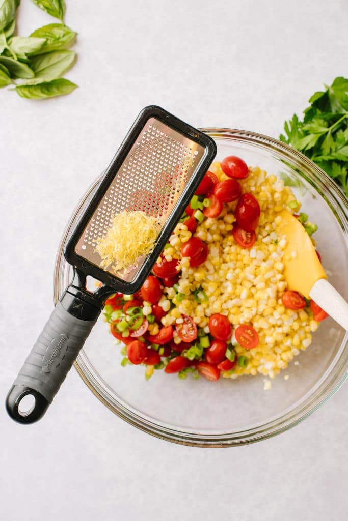 lemon zest on a microplane on side of a bowl of corn, tomatoes, and green onions