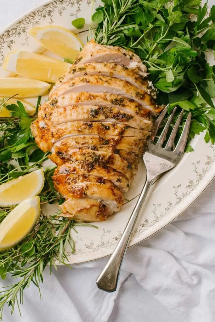 sliced turkey breast on white platter with lemon wedges, fresh herbs and fork