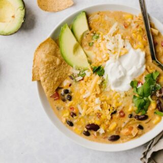 chicken tortilla soup in white bowl with spoon topped with sour cream, cheese, avocado and tortillas on side