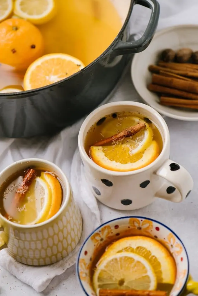 three mugs filled with mulled white wine and garnished with citrus slices with pot of wine in background