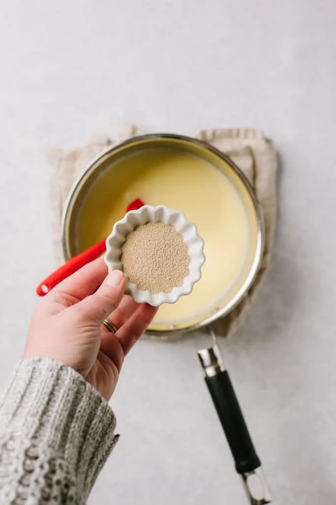 hand holding small bowl of yeast over warm milk and melted butter in pot