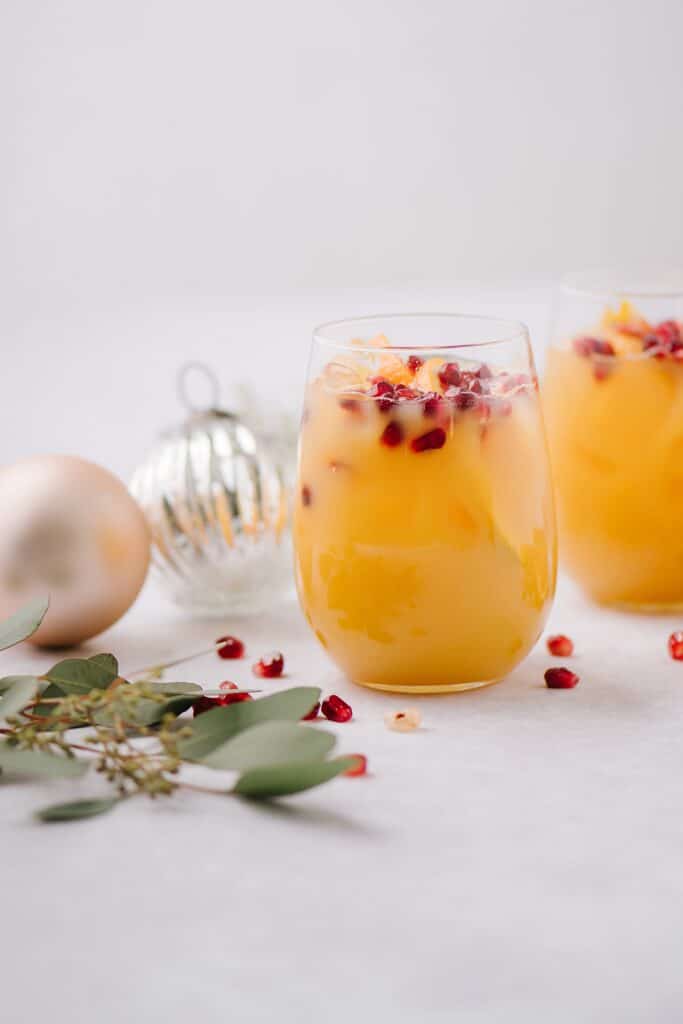 A photo from the side of a Family Stone Sipper cocktail. Pomegranate seeds, holiday ornaments, and greenery, are surrounding the glasses.