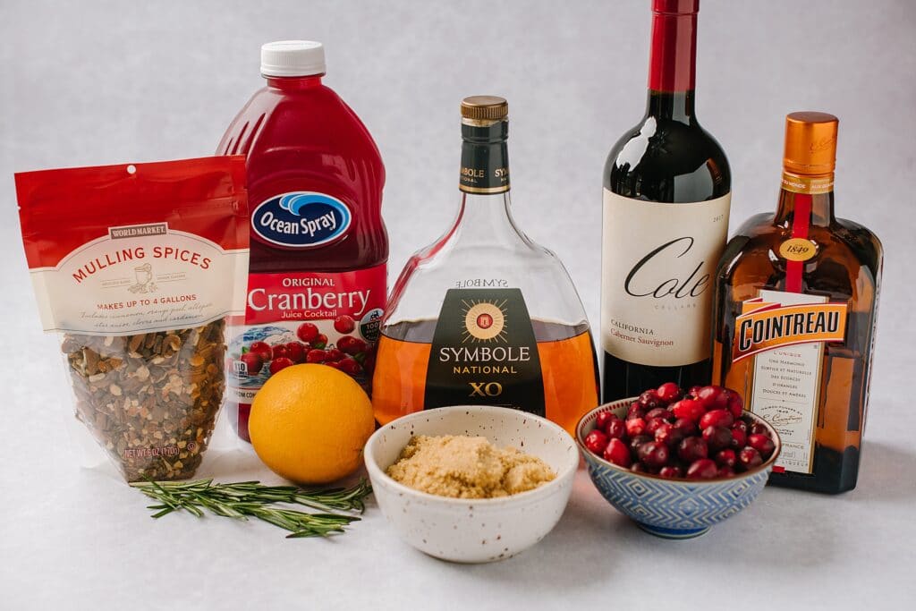 These are the ingredients for the mulled red wine sangria. Mulling spices, cranberry juice, an orange, rosemary sprigs, red wine, Brandy, and Cointreau are lined up in the back. A bowl of brown sugar and a bowl of cranberries are in the foreground.