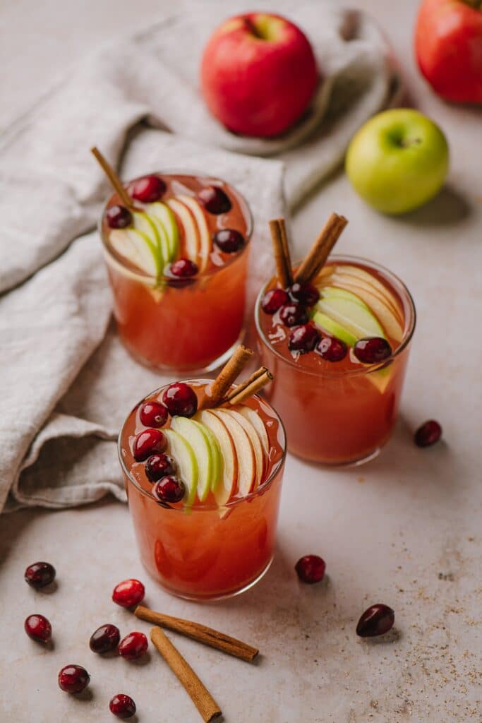 Three glasses of a different version of spiced apple cider cocktails, topped with sliced apple and berries. 