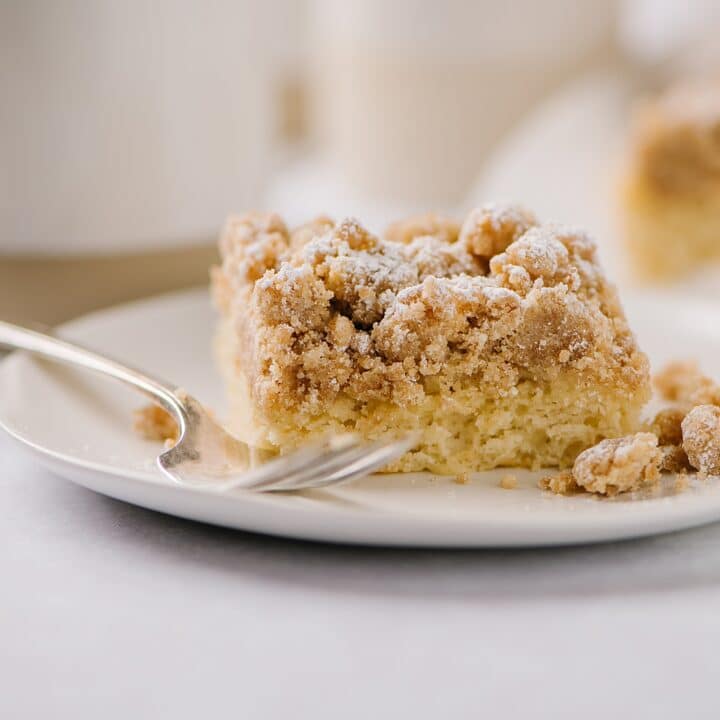 New York Style Crumb Cake on a plate with a fork and a cup of coffee in the background