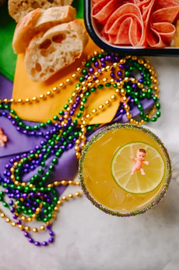 kings cup cocktails with a king cake baby floating on a lime slice with mardi gras beads and yellow green and purple napkins next to it