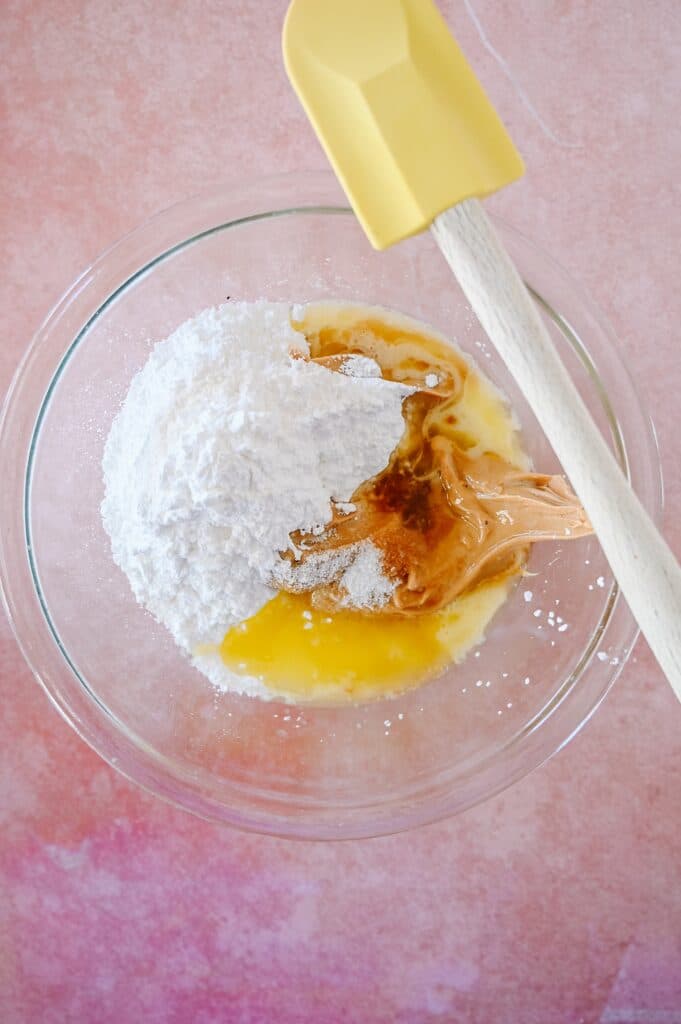 peanut butter, melted butter, powdered sugar, salt, and vanilla in a clear mixing bowl with a yellow spatula on the side