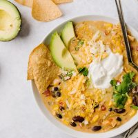 chicken tortilla soup in white bowl with spoon topped with sour cream, cheese, avocado and tortillas on side